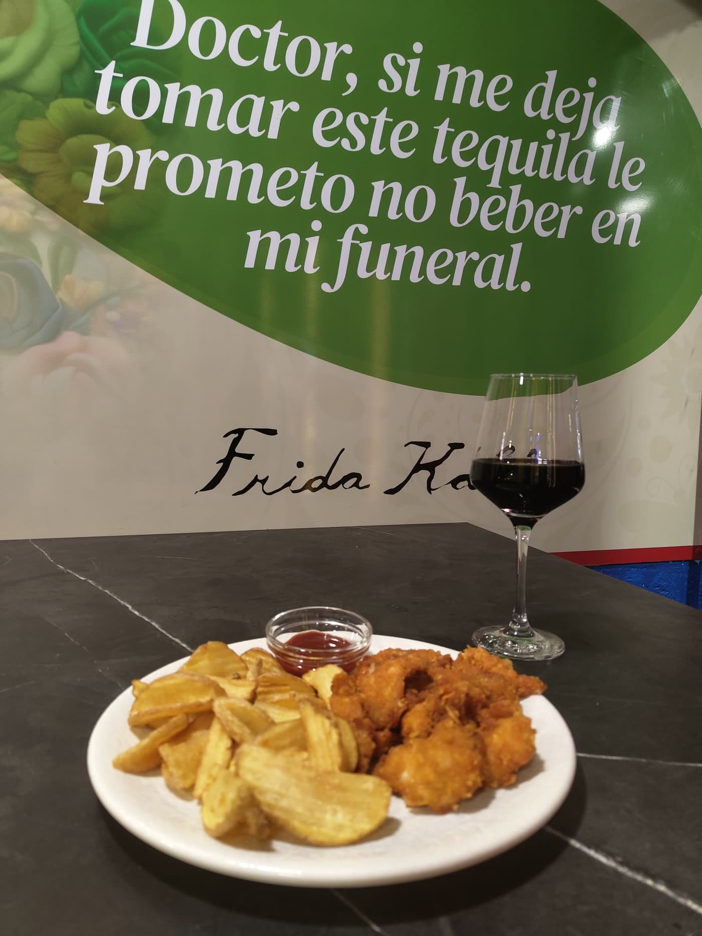 Mural de Frida Kahlo en el local, junto a una copa de vino y un plato de la casa